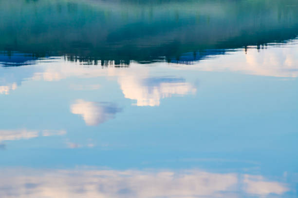 View of a calm thermal pool in Midway Geyser Basin reflecting the dramatic clouds above, with steam drifting across the surface and a dark treeline.
