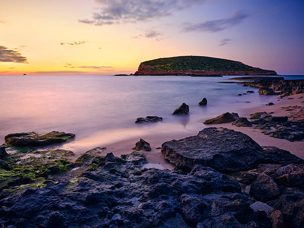 Wide-angle view of one of the most beautiful sandy shores of Platges de Comte (also known as Cala Comte or Cala Conta), a group of scenic beaches located on the western coast of Ibiza. A clear sky, picturesque clouds highlighting the outline of the islet of S'Illa des Bosc, calm waters reflecting the warm, golden light of a Mediterranean sunset, the gentle splashing of the waves on the shore. High level of detail, natural rendition, realistic feel. Developed from RAW.