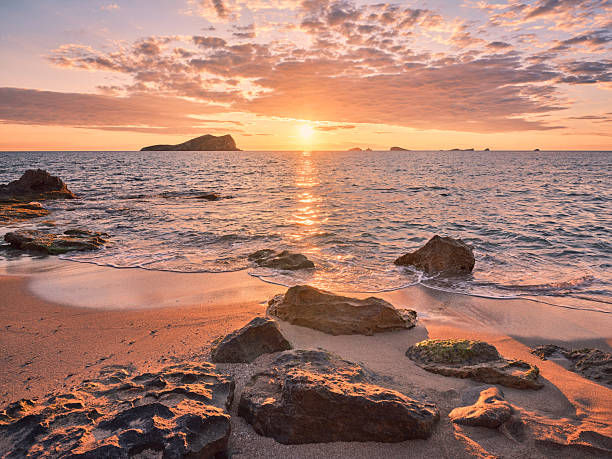 Wide-angle view of one of the most beautiful sandy shores of Platges de Comte (also known as Cala Comte or Cala Conta), a group of scenic beaches located on the western coast of Ibiza. A clear sky, picturesque clouds highlighting the outline of the islets of S'Espartar and Ses Bledes, calm waters reflecting the warm, golden light of a Mediterranean sunset, the gentle splashing of the waves on the shore. High level of detail, natural rendition, realistic feel. Developed from RAW.
