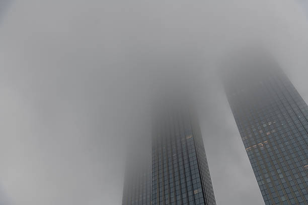 Towering skyscrapers rise above the city, almost disappearing into a thick layer of fog. The mist wraps around their peaks, creating an ethereal and mysterious urban landscape.