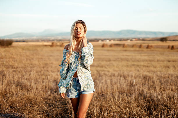 Young woman with blonde hair smiles brightly while standing outdoors in a blue shirt and sunglasses, soaking up the sun on a beautiful day.
