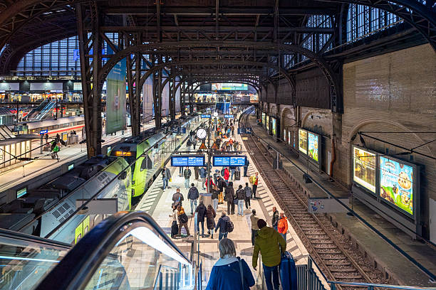 Hamburg, Germany. March 31st, 2025: \nA wide-angle view of the central hall in a busy train station at dusk. The interior features high, metal arches supporting large windows that let in the fading daylight, illuminating the platform below. Several trains are visible on the tracks, and many passengers are walking on the platform or heading towards the escalator. The scene is filled with vibrant lights from signs, advertisements, and the clocks above, adding to the dynamic atmosphere of the station. The bustling crowd and modern infrastructure reflect the energy and functionality of the transportation hub.