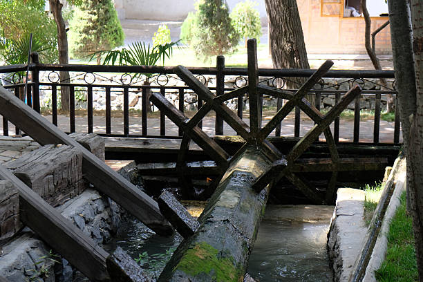 A wooden waterwheel turns gently, drawing water from a small stream in a serene garden. Lush greenery surrounds the area, creating a peaceful atmosphere.