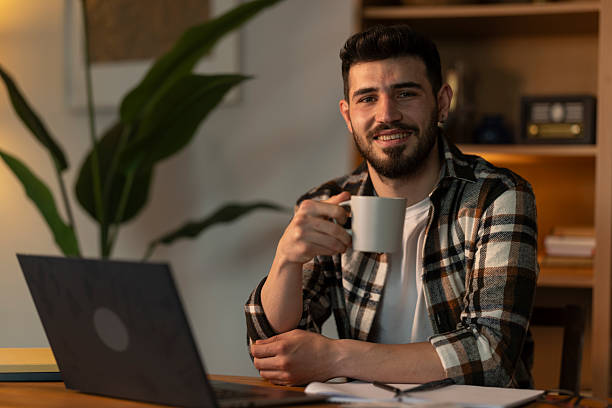 istock Young man smiling and drinking coffee while working from home on his laptop 2208680370