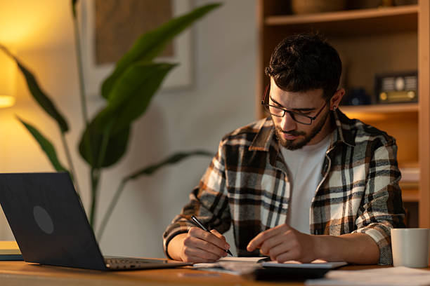 istock Young man sitting at desk writing notes and working on laptop computer at home 2208680371