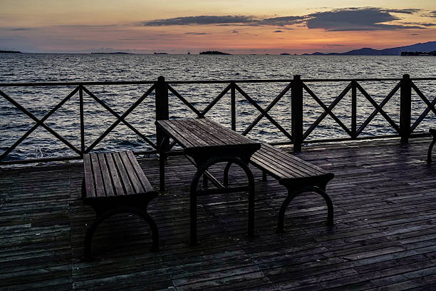 Wooden benches and a table sit on a pier, offering a serene view of the sunset over the aegean sea in izmir, turkey