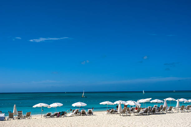 Rows of white beach umbrellas and lounge chairs line the powdery white sands of Grace Bay Beach in Providenciales, Turks and Caicos. Tourists relax and wade into the calm, turquoise waters of the Caribbean under a perfectly blue sky.