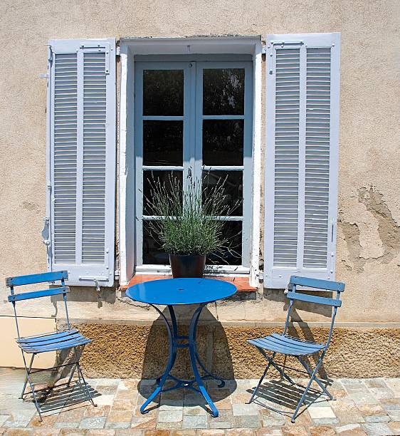 A quaint outdoor setting features two blue chairs and a matching round table placed in front of a window with white shutters. A potted lavender plant adds a touch of greenery to this charming scene on the stone patio.