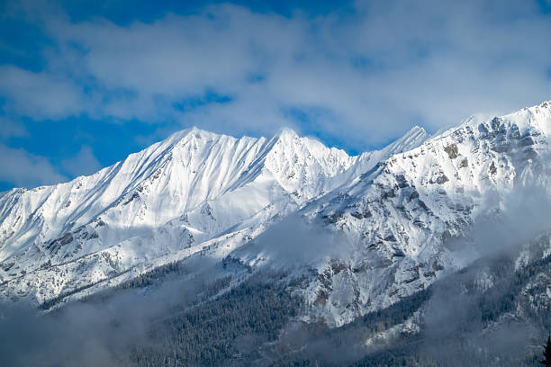 Kootenay National Park snow-covered mountain peaks rise dramatically against a blue sky with scattered clouds. Some trees are visible at the base