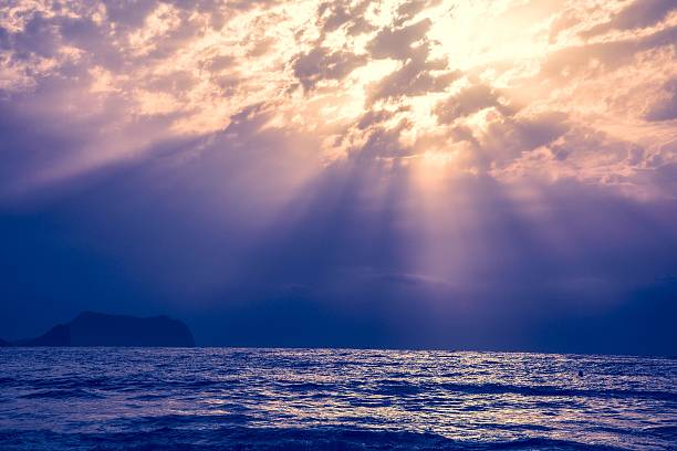 Dramatic seascape with sun rays piercing through clouds over a serene ocean at dusk