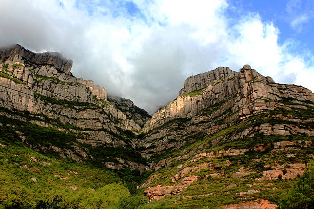 Majestic, layered rock mountains rise dramatically, partially shrouded in clouds, with patches of green vegetation clinging to their steep slopes under a bright sky.