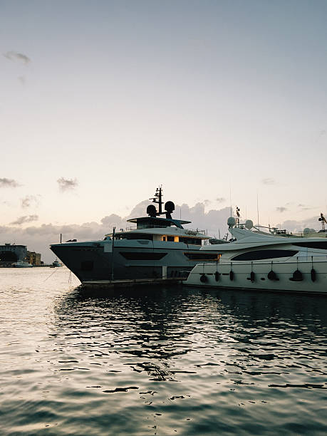 Two large yachts are moored in harbor at sunset, their sleek silhouettes reflected in calm water, creating scene of luxury and tranquility. Luxury yachts moored during golden sunset, maritime ambiance