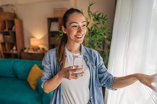istock Young woman drinking water and opening curtains at home 2208793338