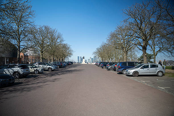 18 March 2025. A tree-lined avenue in Greenwich Park, filled with parked cars, leads to a distant view of Canary Wharf’s skyline, creating a contrast between urban nature and London’s financial district.
