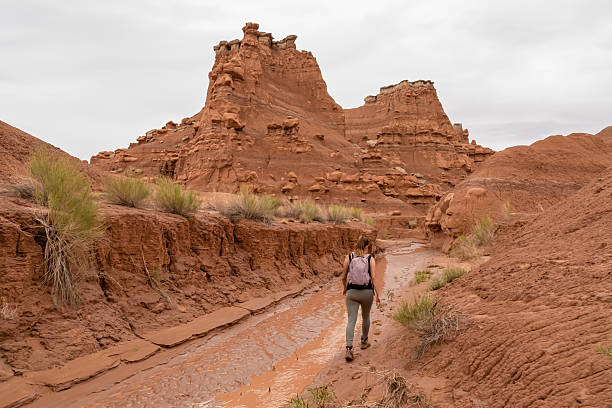 Woman walking along temporary mud river flowing down Goblin Valley State Park, Utah, USA. Scenic canyon hiking trail with unique eroded sandstone hoodoo rock formations called goblins on cloudy day