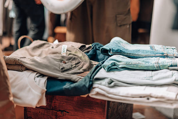 Different folded clothes, including jeans and shirts, laying on a wooden stall at a flea market, with a price tag visible and blurred people in the background