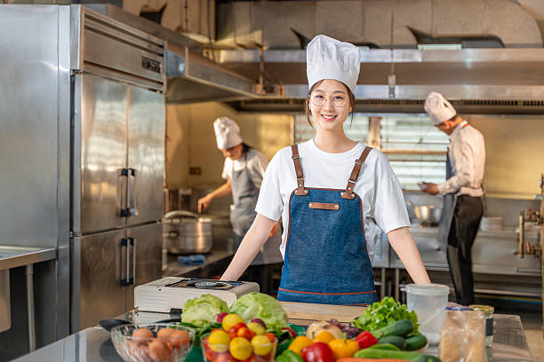 istock Happy young Asian female chef standing in her kitchen confidently smiling at camera while two other chefs working in the background 2208806508