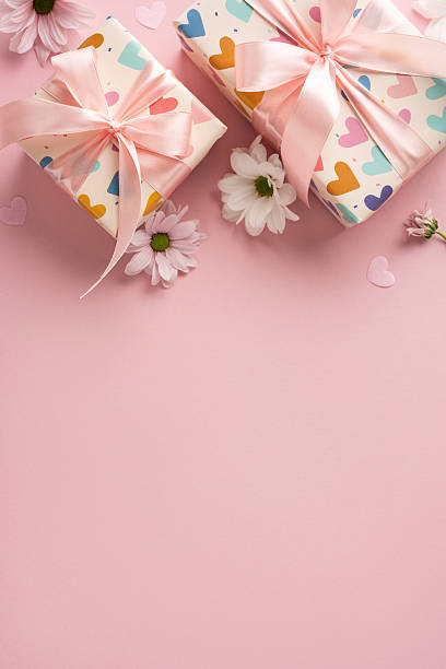 Beautifully wrapped vertical gift boxes tied with pink ribbons, surrounded by white daisies and heart accents, placed on a pink surface