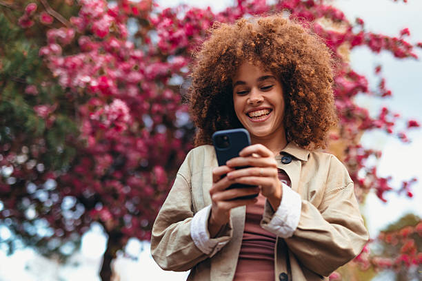 istock Young woman using smartphone and smiling near blooming tree 2208811132