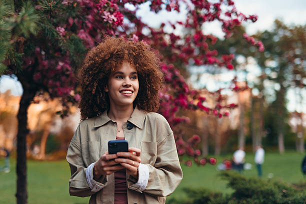 istock Young woman using smartphone in a park with blooming trees 2208811200