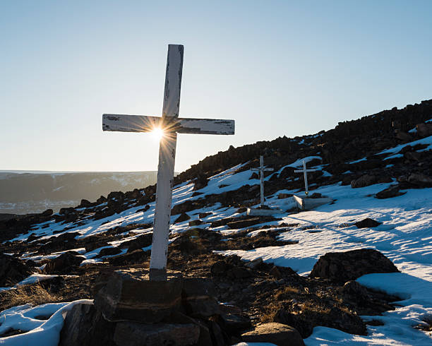 Snow blankets the landscape as sunset casts a warm glow through the crosses in a serene Greenland churchyard. The scene evokes peace and contemplation amid the natural beauty.