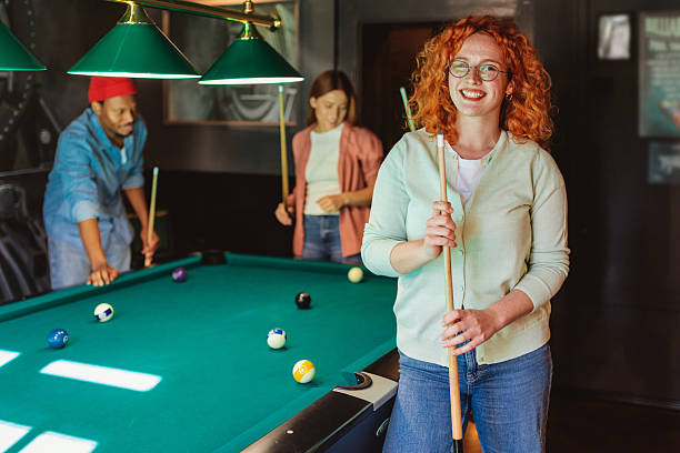 A Vibrant Young Woman With Striking Red Hair Enjoys a Night Out at a Billiard Club, Looking Directly at the Camera While Her Friends Engage in a Game Behind Her. The Friendly and Social Setting Makes This Stock Photo Ideal for Bar and Pub Advertisements, Fashion Brands Targeting Casual Nightlife Style, and Editorial Content on Fun Social Activities.