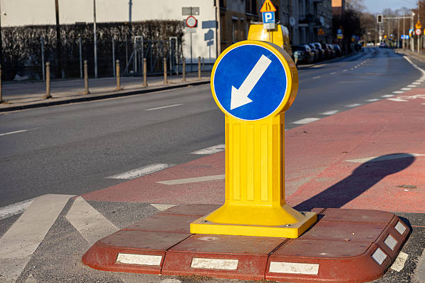 istock Directional road sign with white arrow on yellow post indicating traffic must pass on the left, standing on pedestrian island in urban street under sunlight 2208844680