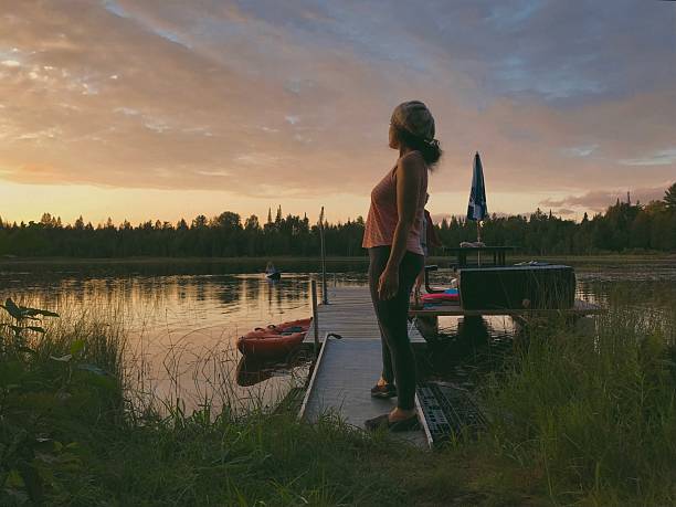 A rich, moody sunset scene on a small Wisconsin lake, captured from a low angle through lakeside grass. A middle-aged woman in a beret stands confidently on a wooden dock, while a child kayaks in the background. A grandmother relaxes on a homemade pontoon-style float boat featuring picnic tables and an umbrella, tied to the dock. The setting sun casts golden light across the water and surrounding forest, creating a warm yet cinematic atmosphere of connection, nature, and generational bonds. Taken on Cotas Lake in Pembine, Wisconsin.