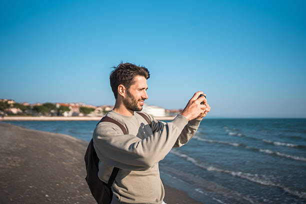 Candid portrait of young Caucasian male in beige sweater photographing with smartphone on sandy beach, against backdrop of rolling waves and clear blue horizon.