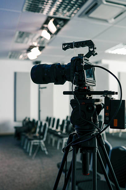 istock Professional video camera on a tripod set up in a modern conference room, ready for filming or live streaming, with rows of empty chairs and ceiling lights in the background 2208858518