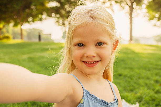 A blonde girl joyfully takes a selfie while in a green, sunny park. The image exudes happiness and showcases the beauty of childhood. Soft sunlight filters through the trees, enhancing the warm and inviting atmosphere.