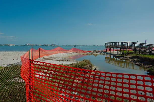 Beach front Renourishment Area With Red Fence and Pedestrian Bridge. A coastal environment featuring a red safety fence by the water, shielding restored land, with a wooden footbridge under a clear blue sky, demonstrating environmental restoration and safety measures. View out over Boca Ciega Bay Gulfport, Florida gulfport florida photos stock pictures, royalty-free photos & images