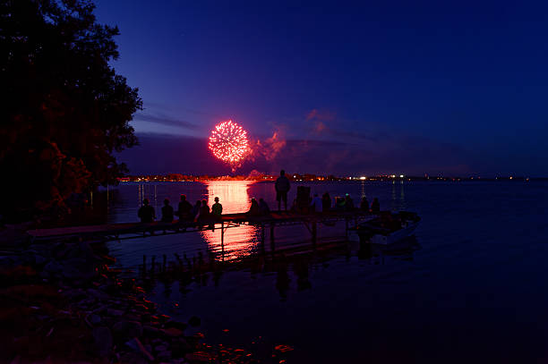 Canada Day fireworks light up the Bay of Quinte, Ontario, sky in vibrant red. Silhouettes of people on a wooden dock watch as the fireworks' reflection shimmers on the calm water.