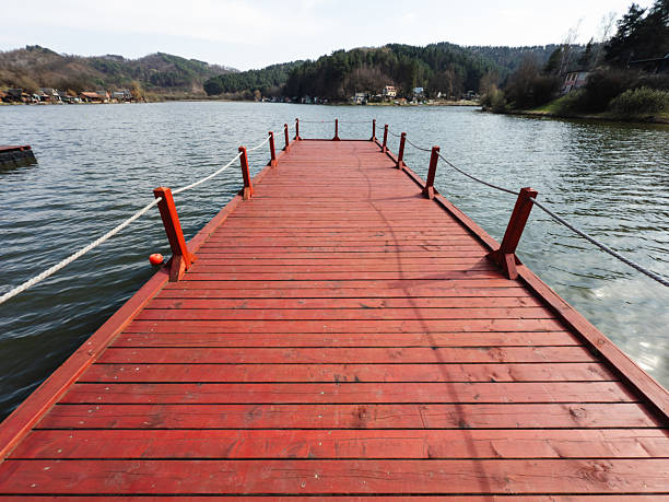 A vivid red wooden pier stretches out into a tranquil lake, framed by distant forested hills and scattered lakeside cottages. The still water and soft lighting create a peaceful and contemplative mood, while the central perspective leads the viewer’s eye toward the horizon. The scene captures a serene moment in nature.