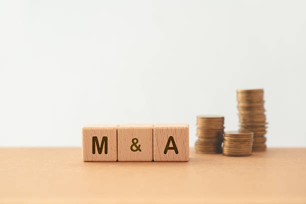 istock M and A, Mergers and Acquisitions, text on wooden cube blocks with blurred stack of coins  including copy space 2208912418