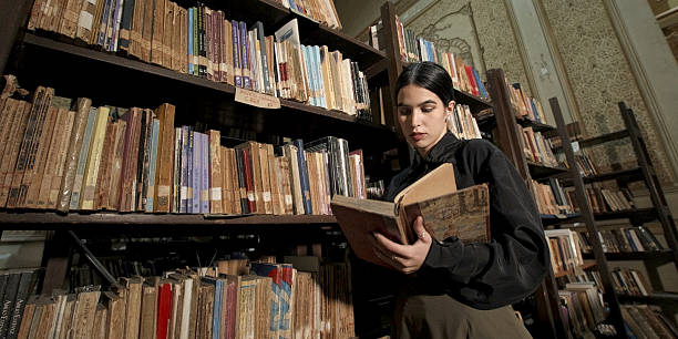 Young woman reading an old book while standing in a vintage library, surrounded by bookshelves filled with classic literature