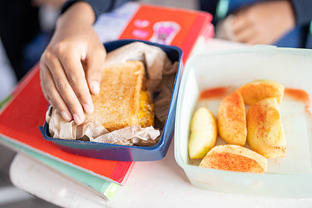 A group of middle schoolers sit side by side in a bright classroom, eating homemade meals from compartmented lunch boxes and metal tiffins; water bottles, chapati, rice, and snacks are spread across the desks as they chat and unwind, capturing the everyday warmth of school community and midday routines.