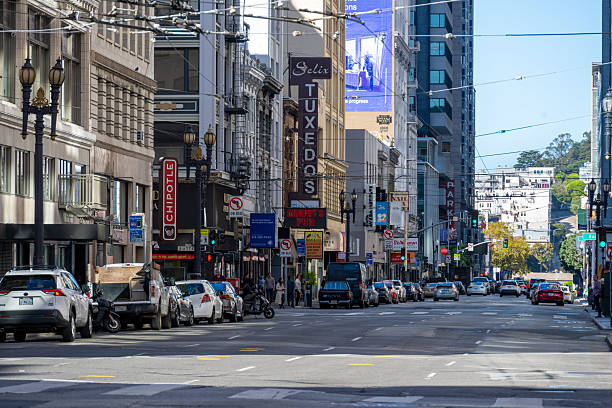 A typical sunny day in downtown San Francisco, California, captured from a street-level perspective. Cars are parked along the road, pedestrians are crossing the street, and various commercial signs like tuxedo shops, Stripe ads, and traffic signs give the image an authentic urban vibe. The tall buildings cast soft shadows along the street, creating a vibrant, lived-in city atmosphere.