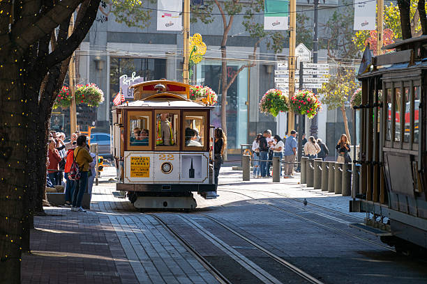 A traditional San Francisco cable car on Powell Street, surrounded by locals and tourists waiting to board. This vibrant city scene captures one of the most iconic modes of transport in the city, evoking nostalgia and charm. The sunlit atmosphere, hanging flower baskets, and urban energy showcase the timeless appeal of downtown San Francisco.