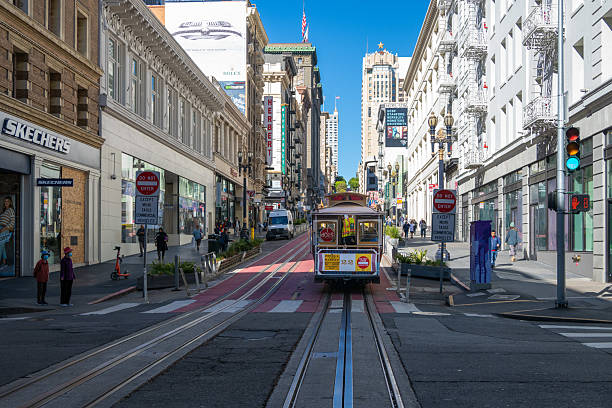 A traditional San Francisco cable car on Powell Street, surrounded by locals and tourists waiting to board. This vibrant city scene captures one of the most iconic modes of transport in the city, evoking nostalgia and charm. The sunlit atmosphere, hanging flower baskets, and urban energy showcase the timeless appeal of downtown San Francisco.