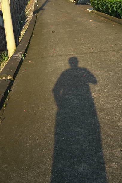 A solitary figure's elongated shadow leads the way up a steep, sunlit road, symbolizing a personal journey, aspiration, or the path ahead. Captured from a first-person viewpoint, the image evokes determination, progress, and the introspective feeling of moving forward alone towards a goal under the morning/afternoon sun.
