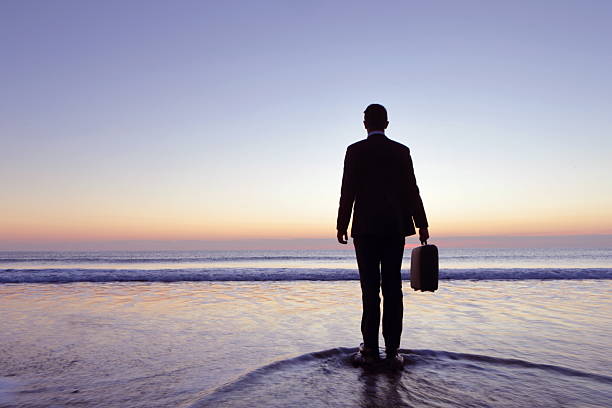 Businessman stands in sea shallows, sunset, Cornwall