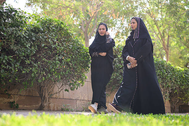 Low angle view of Middle Eastern women chatting and walking side by side in abayas. Two hijabi friends enjoying a bright day outdoors, surrounded by lush greenery.