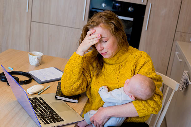 Young mother in yellow sweater working on laptop while holding baby in modern kitchen with notebooks, coffee cup, headphones, and kitchenware.