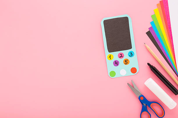 istock Smartphone, glue, scissors, marker, pencil and colorful application paper on light pink table background. Pastel color. Creating child gadget for playing. Closeup. Empty place for text. Top down view. 2208944462