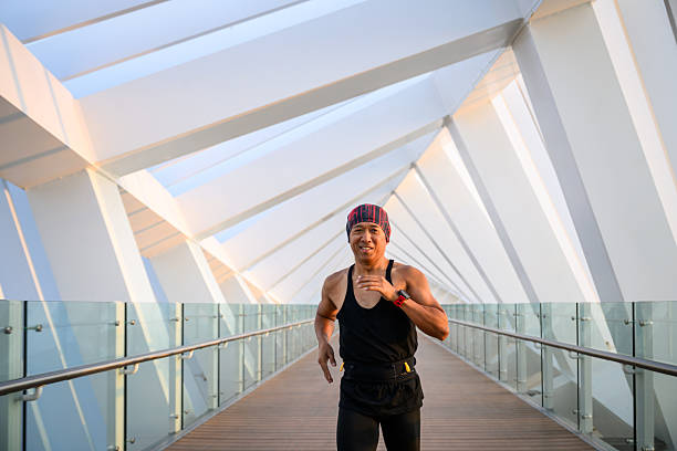 Three length shot of a happy Asian man jogging towards the camera on a pedestrian bridge with geometric architectural detail. Front view of a runner working out in the city.