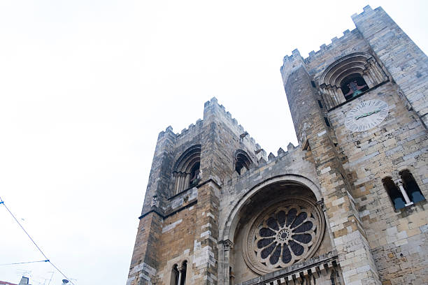 Lisbon cathedral, also known as se de lisboa, stands tall against a cloudy sky, showcasing its intricate rose window, clock, and fortified architecture, a blend of romanesque and gothic styles