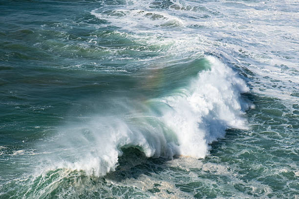 Massive ocean wave crashing near rocky coastline, generating dramatic water spray with colorful rainbow arcing above turbulent seascape