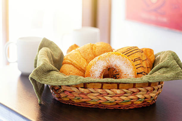 Wicker basket overflowing with fresh pastries, including croissants and a sugared donut, resting on a dark wooden table beside a white mug in soft focus