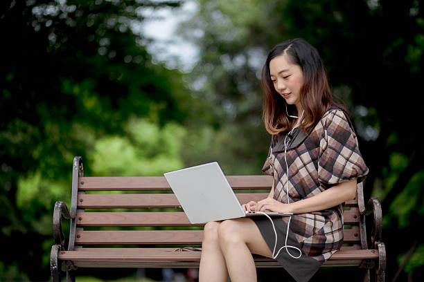 istock Smiling young woman using laptop on a park bench with earphones on, enjoying remote work outdoors surrounded by trees and peaceful green nature. 2208964239
